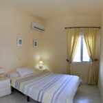 A cosy bedroom with a double bed, striped bedding, two white bedside tables with lamps, a window with sheer yellow curtains, framed pictures on the wall, and an air conditioner above the bed.