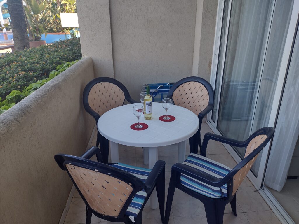 A small round table with four chairs on a balcony, featuring a bottle of white wine and two glasses. Sunlight shines on the scene, with greenery and a resort or hotel visible in the background.