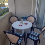 A small round table with four chairs on a balcony, featuring a bottle of white wine and two glasses. Sunlight shines on the scene, with greenery and a resort or hotel visible in the background.
