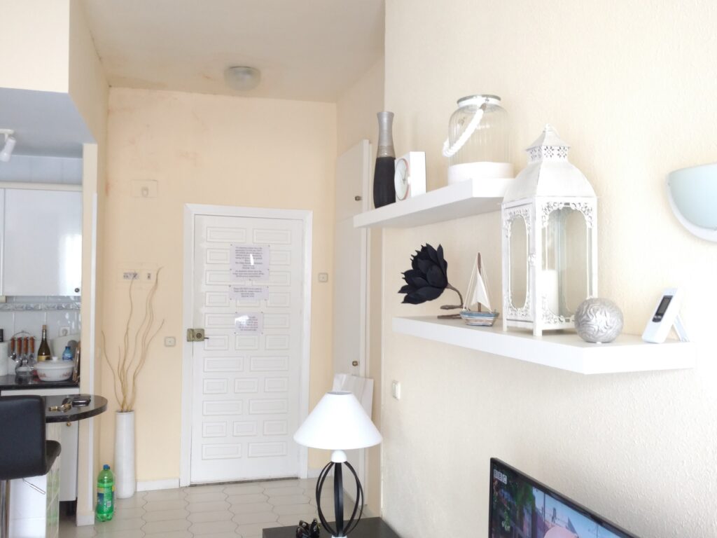 A bright living room with white walls, shelves displaying decorative items like a lantern, vase, and sculptures. A white door and lamp are visible, with part of a kitchen and a green bottle in the corner.