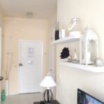 A bright living room with white walls, shelves displaying decorative items like a lantern, vase, and sculptures. A white door and lamp are visible, with part of a kitchen and a green bottle in the corner.