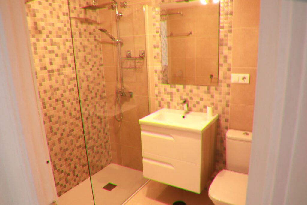 Modern bathroom with a glass shower enclosure, wall-mounted shower fittings, a white sink unit with drawers, a large mirror, mosaic tile accents, and a white toilet.