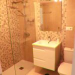 Modern bathroom with a glass shower enclosure, wall-mounted shower fittings, a white sink unit with drawers, a large mirror, mosaic tile accents, and a white toilet.