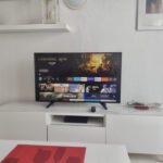 A flat-screen TV on a white cupboard displays the Carnival Row show selection screen. The cupboard holds various items, including a remote control and decorative objects, in a bright, modern living room.