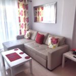 Bright living room with a beige corner sofa, red and floral cushions, a white coffee table with red accents, floral curtains, wall mirror, side table with a hat and games controller, and large windows with sheer curtains.
