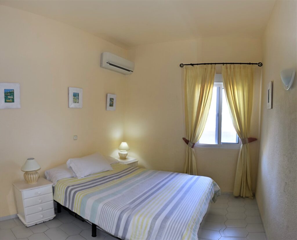 A simple bedroom with a double bed, striped bedding, two white bedside tables with lamps, three pictures on the wall, a window with sheer yellow curtains, and an air conditioner above the bed.
