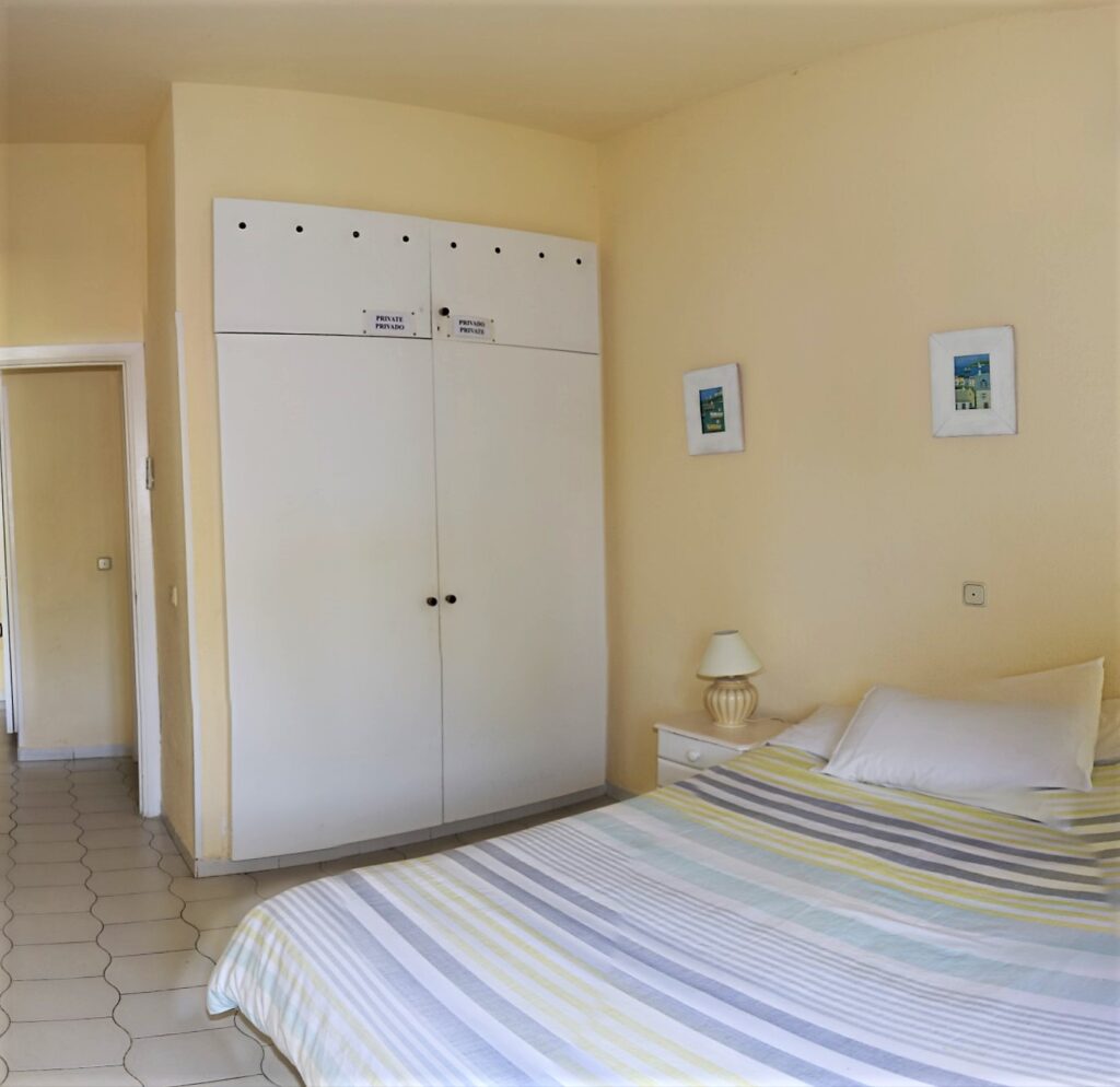 A tidy bedroom with pale yellow walls, a double bed with a striped duvet, a white wardrobe with labels, a small bedside table with a lamp, and two framed pictures hanging above the bed.