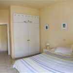 A tidy bedroom with pale yellow walls, a double bed with a striped duvet, a white wardrobe with labels, a small bedside table with a lamp, and two framed pictures hanging above the bed.